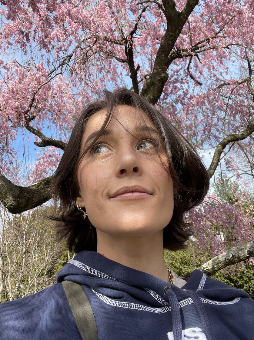 Em is a white woman with a straight brown bob who is here standing in front of a blossoming cherry tree. She is wearing a blue hoodie.