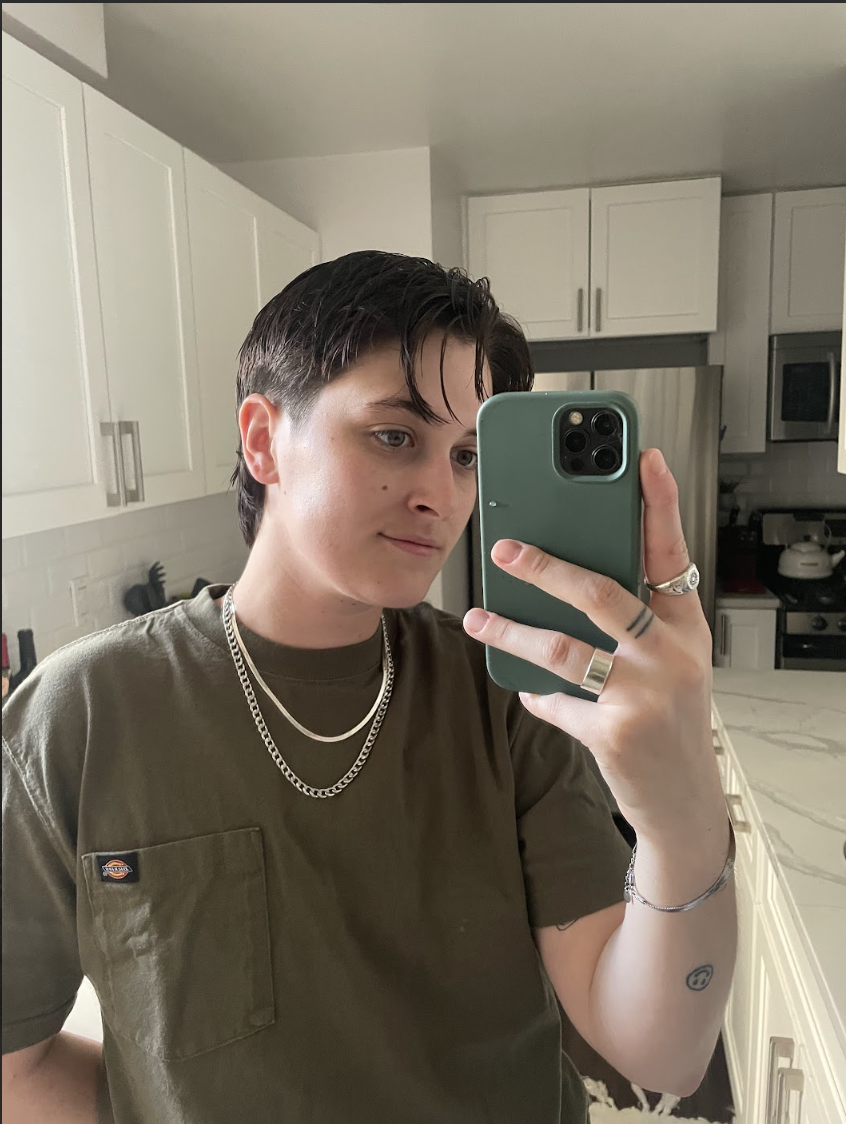 Motti is a white trans masc guy with short brown hair. They are here taking a selfie with phone visible while in a kitchen. They're wearing a sage green tee and multiple gold chains.