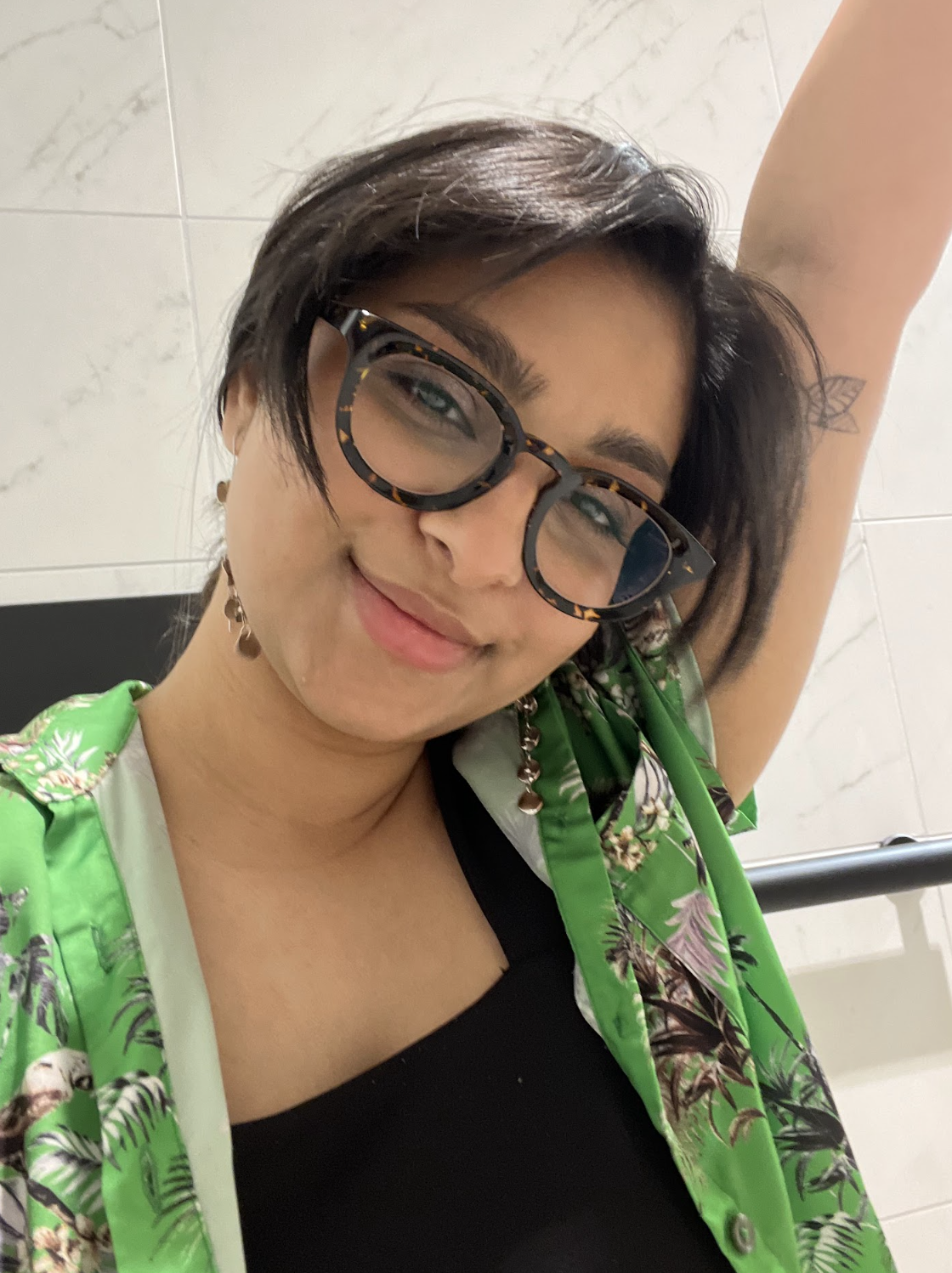 Ashni, a South Asian person, is raising an arm in this photo and smiling playfully. She has short straight dark hair and glasses and her shirt is a colorful button up.