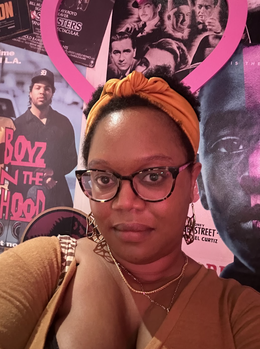 Carmen is a Black woman with short curly hair done up in an orange headband and glasses. She is looing at the camera and standig in front of a wall that is covered in movie posters. She is wearing a burnt marigold shirt and big dangly earrings.