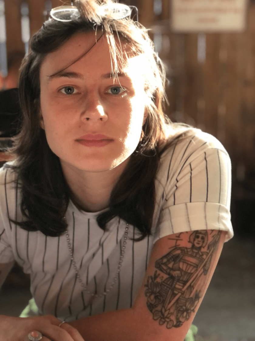 a photo of Gabbie who is a white woman who here is leaning forward and looking at the camera. she has a striped shirt on and a tattoo visible on her arm. she has her glasses pushed up onto her head and has medium length straight brown hair.