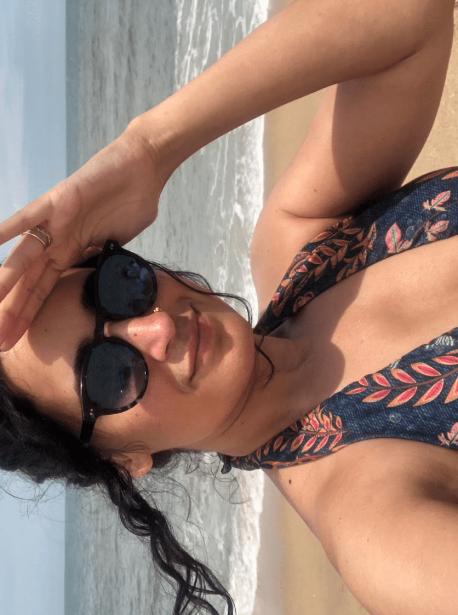 Kayla, a South Asian woman wears a bathing suit and shields her eyes from the sun while wearing sunglasses on a sandy beach with ocean behind her. She has dark hair that is up.