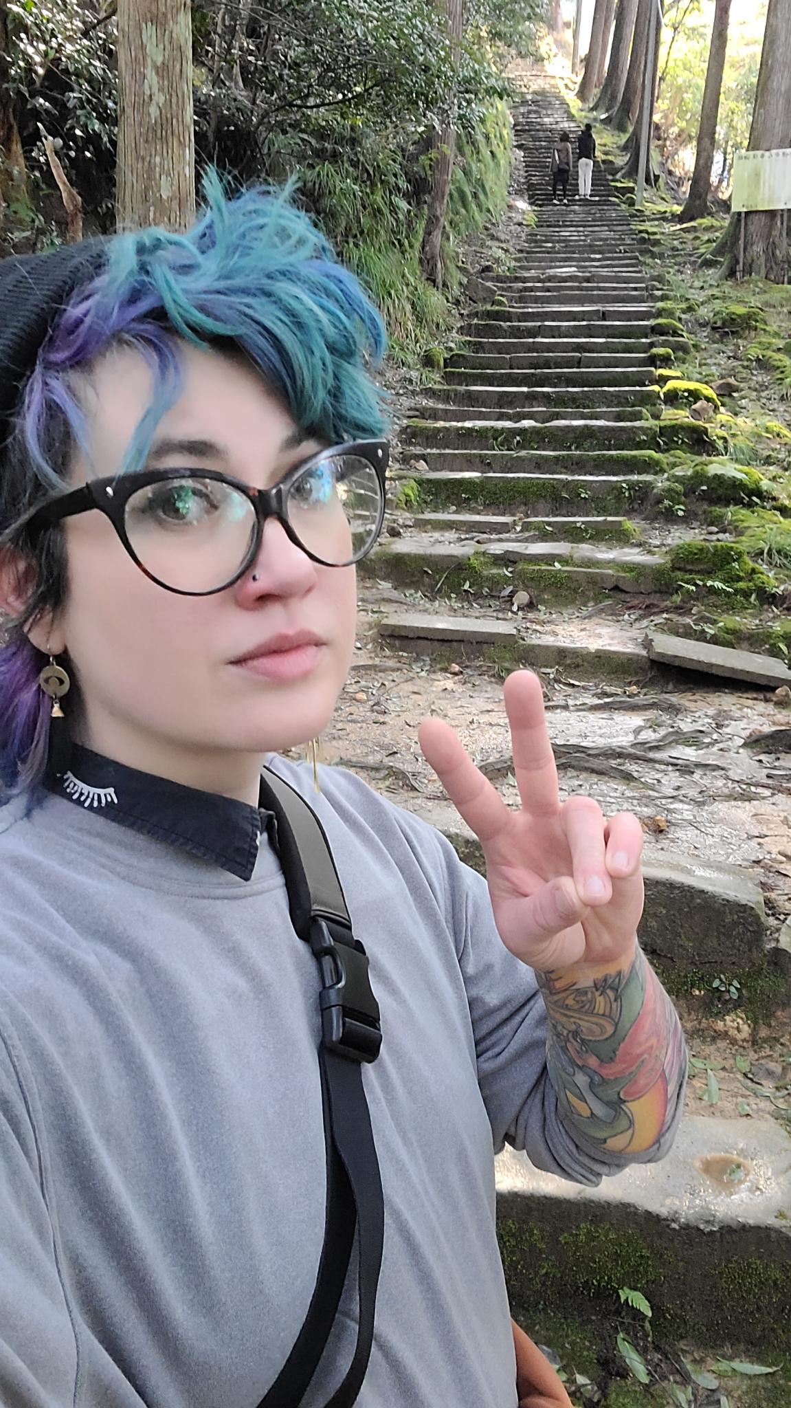 a photo of Nico who is a white genderfluid person. Nico is wearing a collared shirt and gray pullover and has blue and purple hair and glasses. They are making a peace sign and standing in front of a long line of stone stairs in a forest