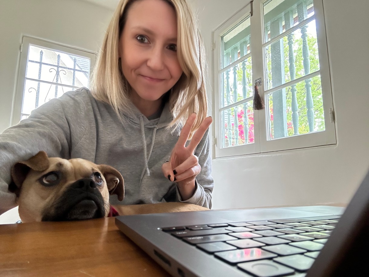 A photo of Riese a white agender person making a peace sign while seated at a computer. There is a wrinkly dog in their lap and a sunny white room behind. Riese has shoulder length blonde hair.