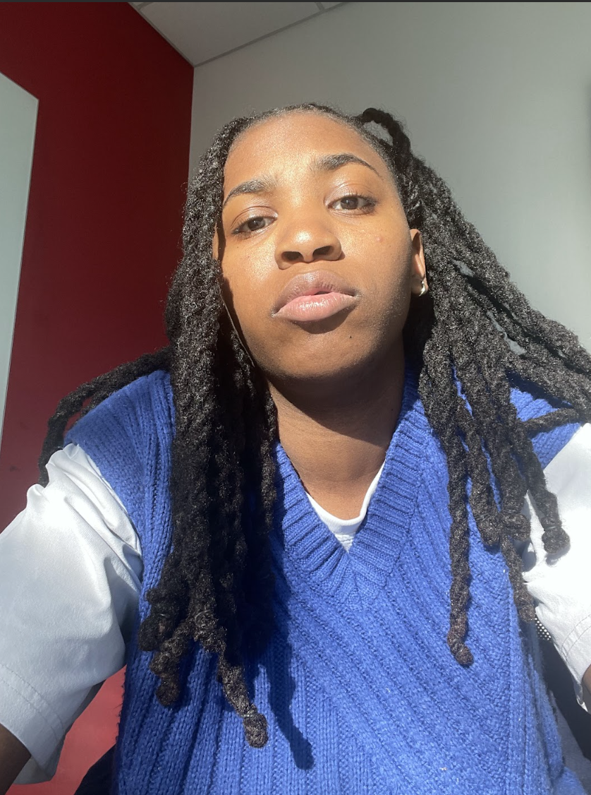 A photo of Tima a Black woman with long locs wearing a tee shirt and sweater vest looks down at the camera