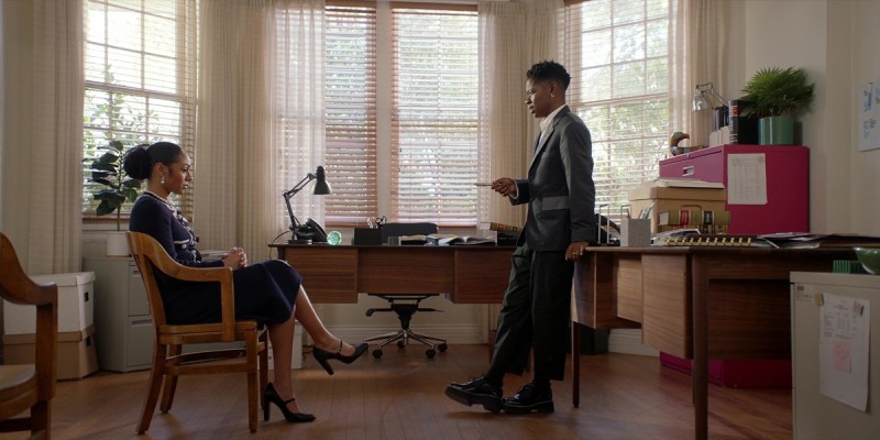 Coop leans on a desk, while Patience sits across from her, in a chair, participating in witness prep.