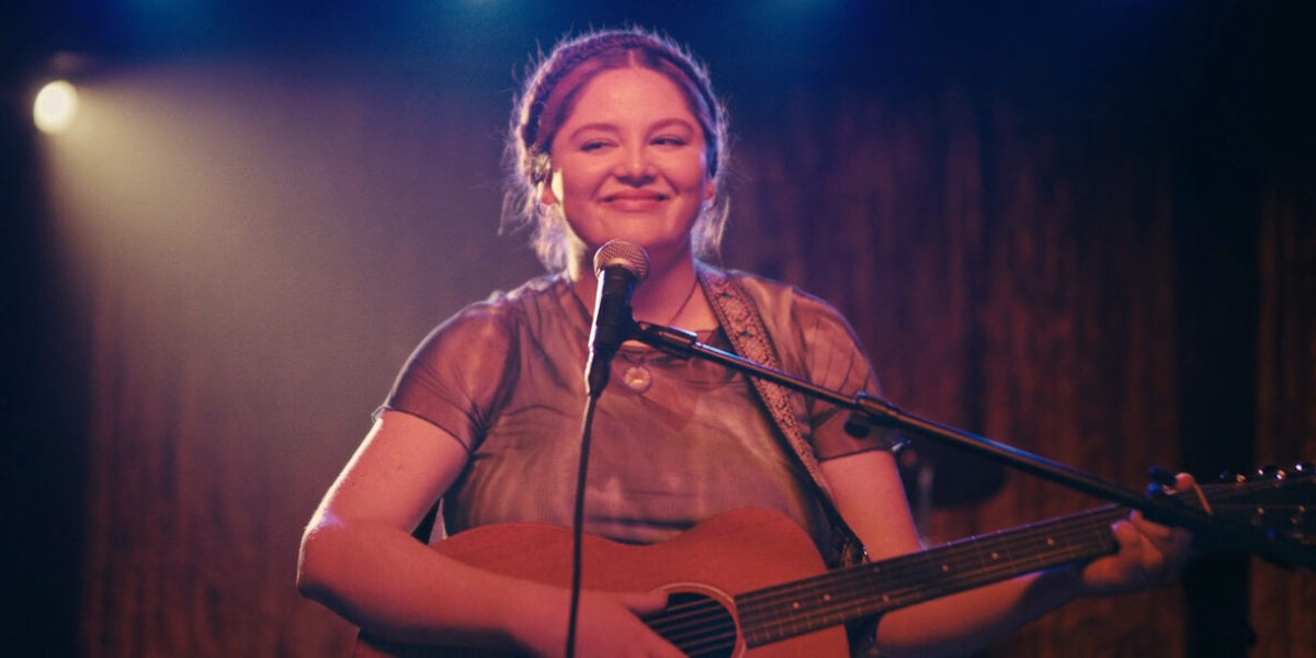 Cora Bora: Meg Stalter stands at a microphone with an acoustic guitar