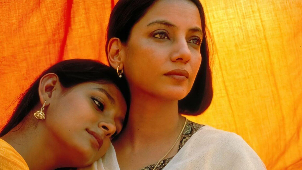 A woman rests her head on another woman's shoulder with an orange background.