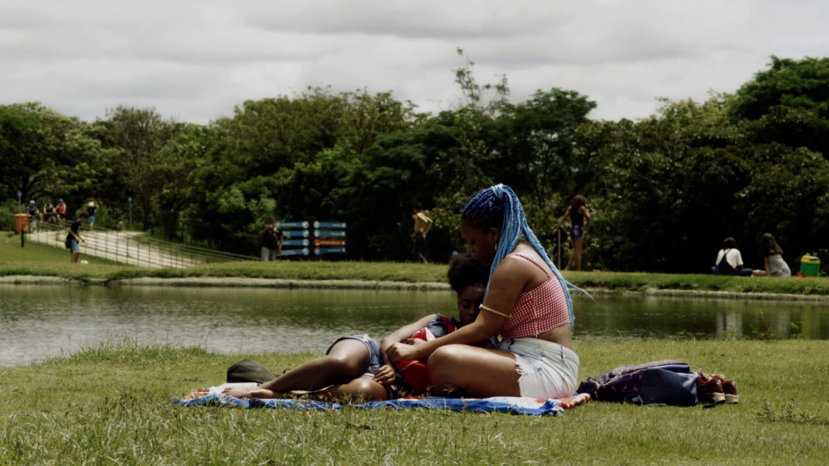 Two Black women cuddle on a picnic blanket in a park.