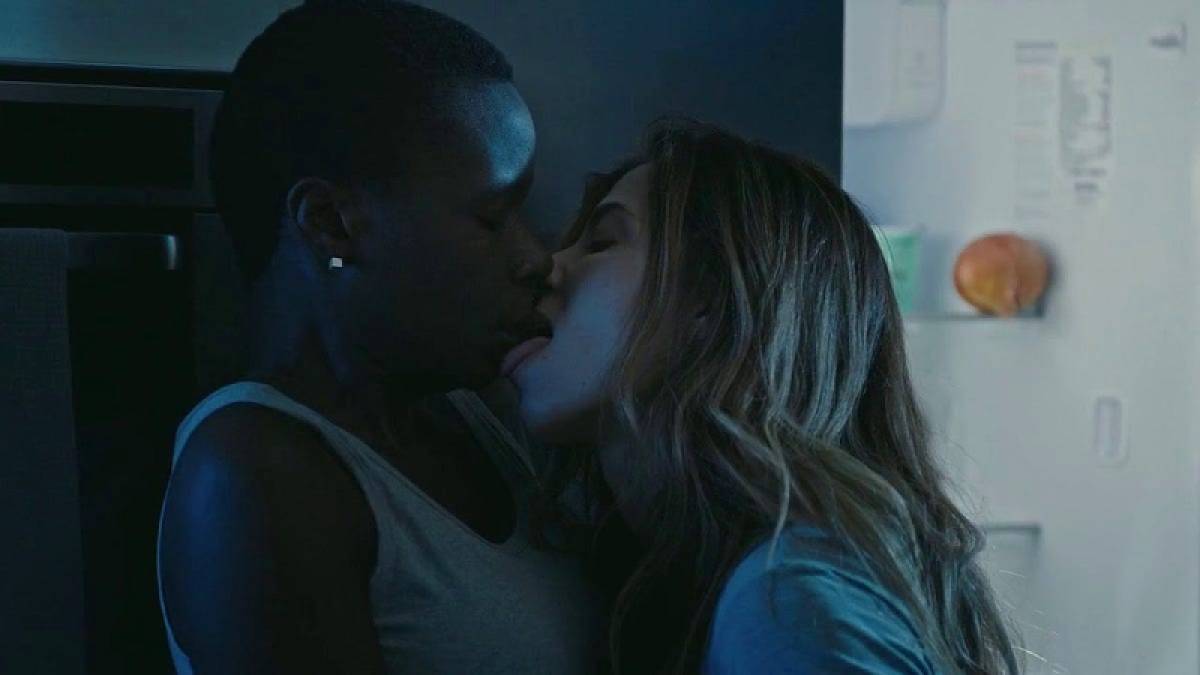 A white woman with long hair leans forward and licks the mouth of a Black woman with short hair who leans away.