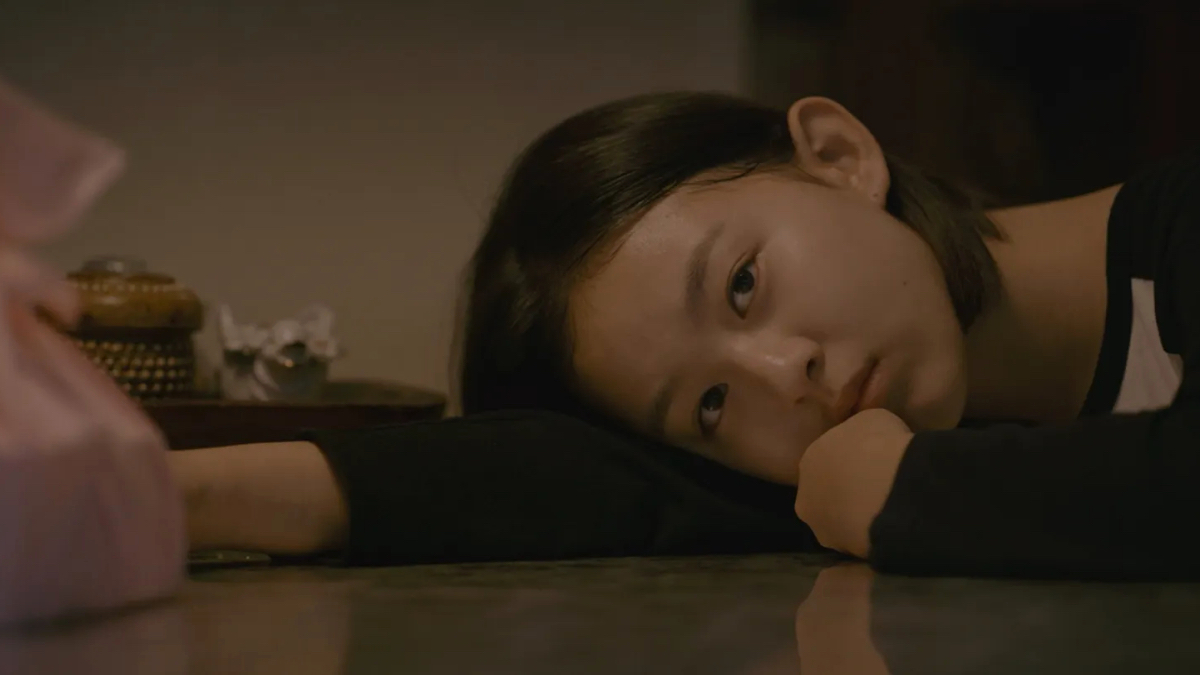 A young girl lies her head on a table with her arm outstretched and looks toward the camera.