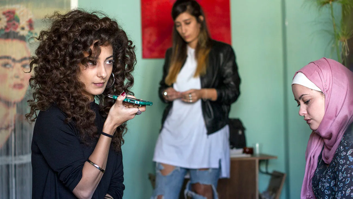 A woman with big curly hair talks on the phone while another woman texts standing in the background and a third woman in a hijab looks on.