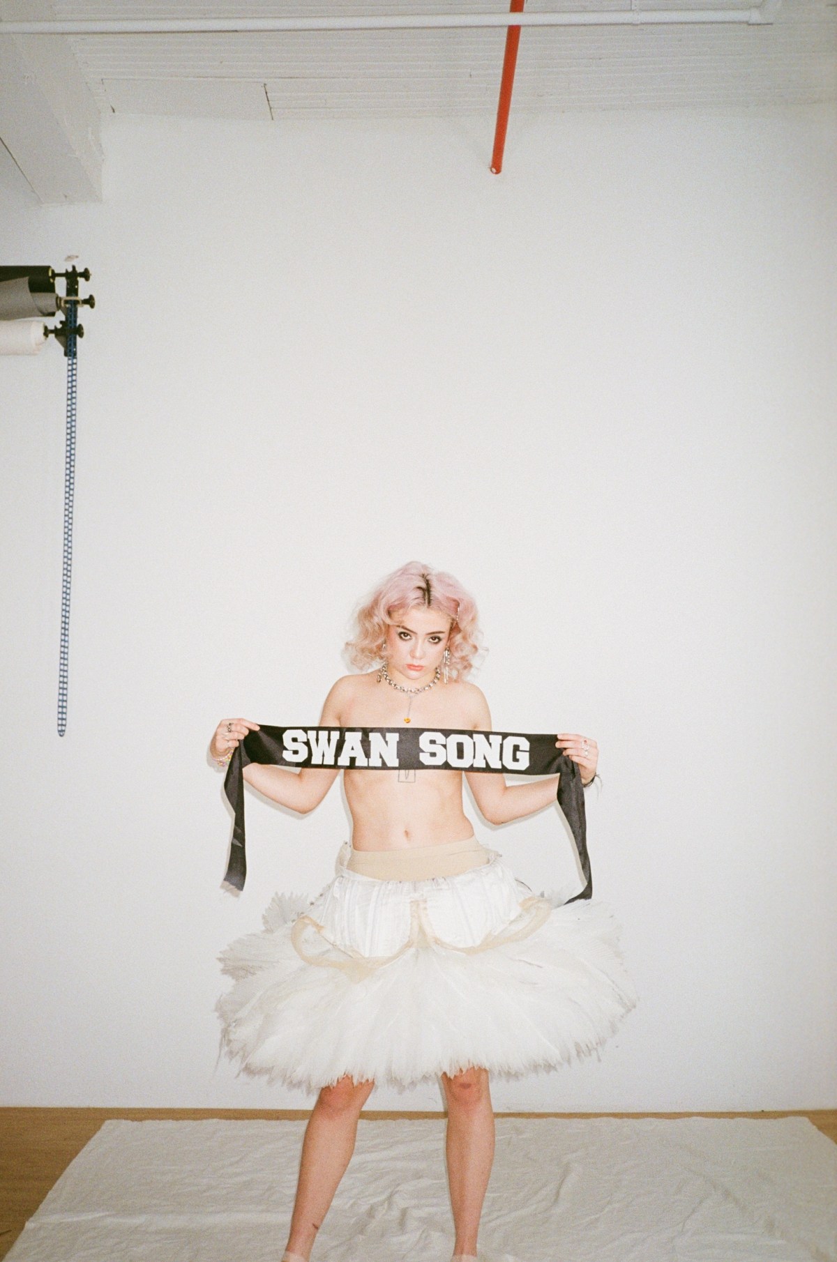 Queer ballerina Shaelynn Estrada topless in a tutu holds up a black banner that says Swan Song