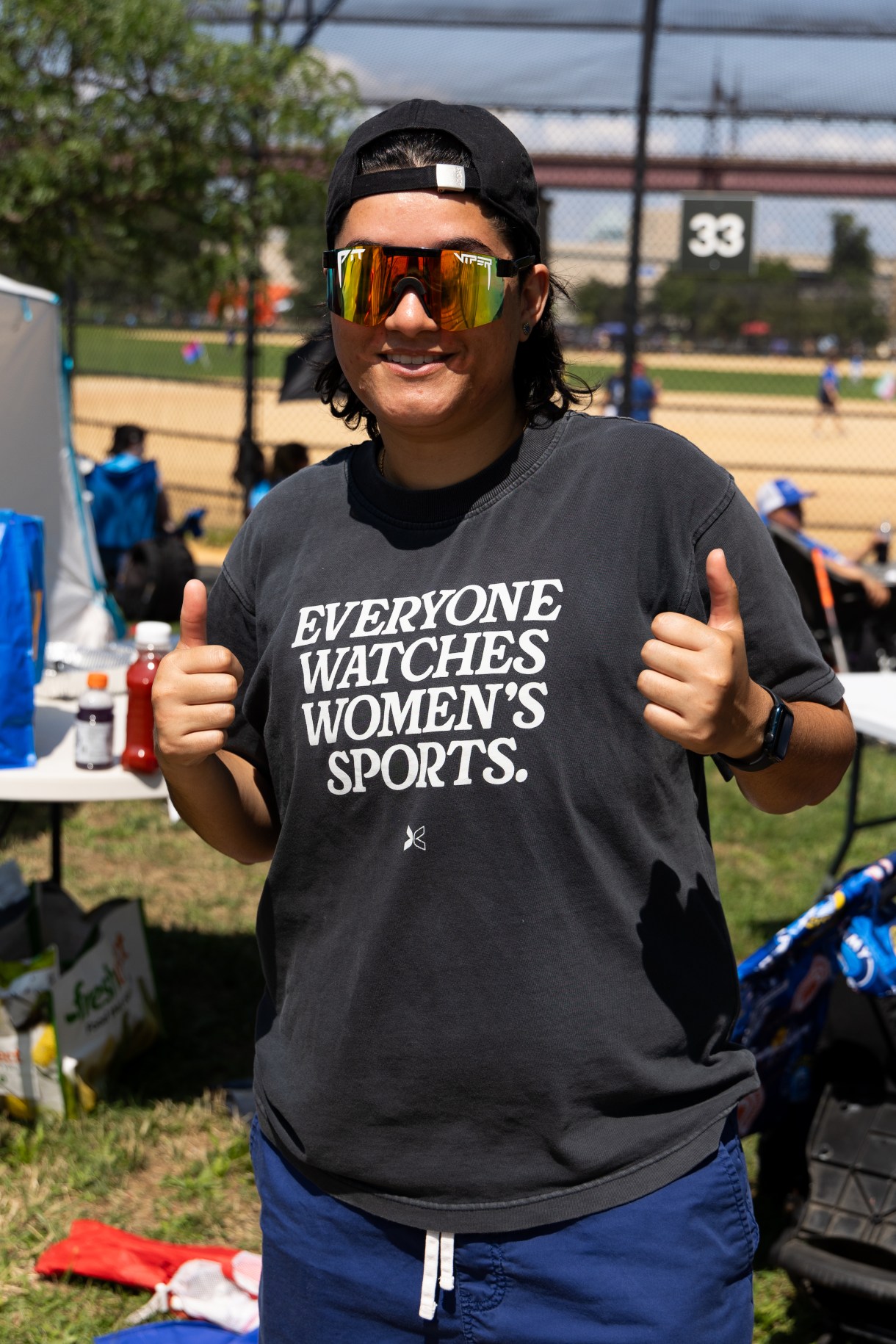 a person flashes thumbs up while wearing a shirt that says EVERYONE WATCHES WOMEN'S SPORTS