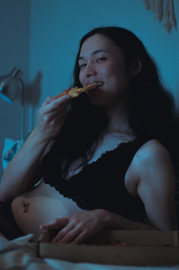 a person eats pizza in bed in a black bra while smiling at camera