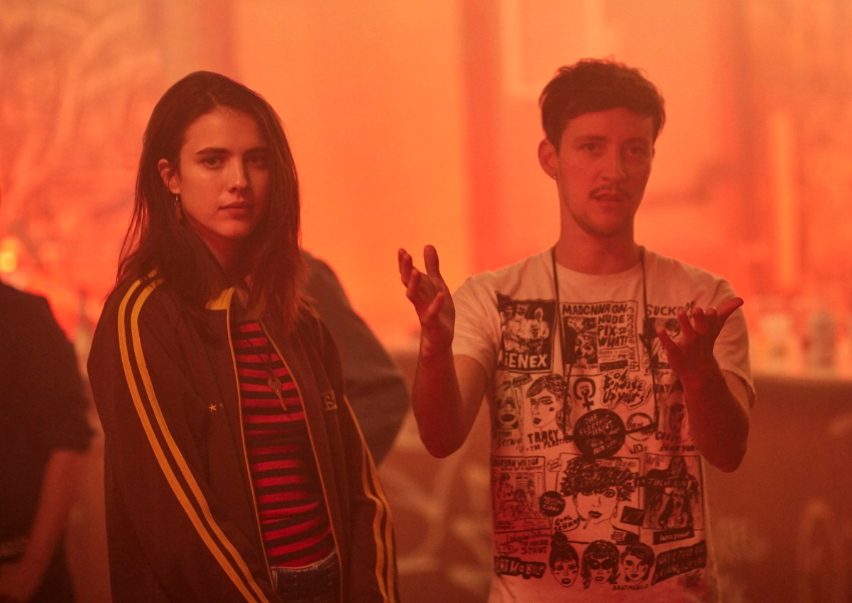 Rhys talks with his hands standing next to Margaret Qualley in orange light