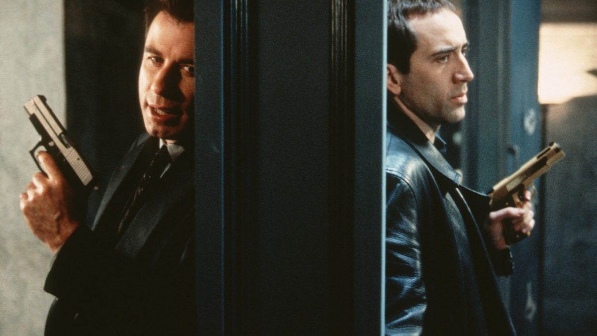 John Travolta and Nicolas Cage hold guns with a wall in between them back to back.