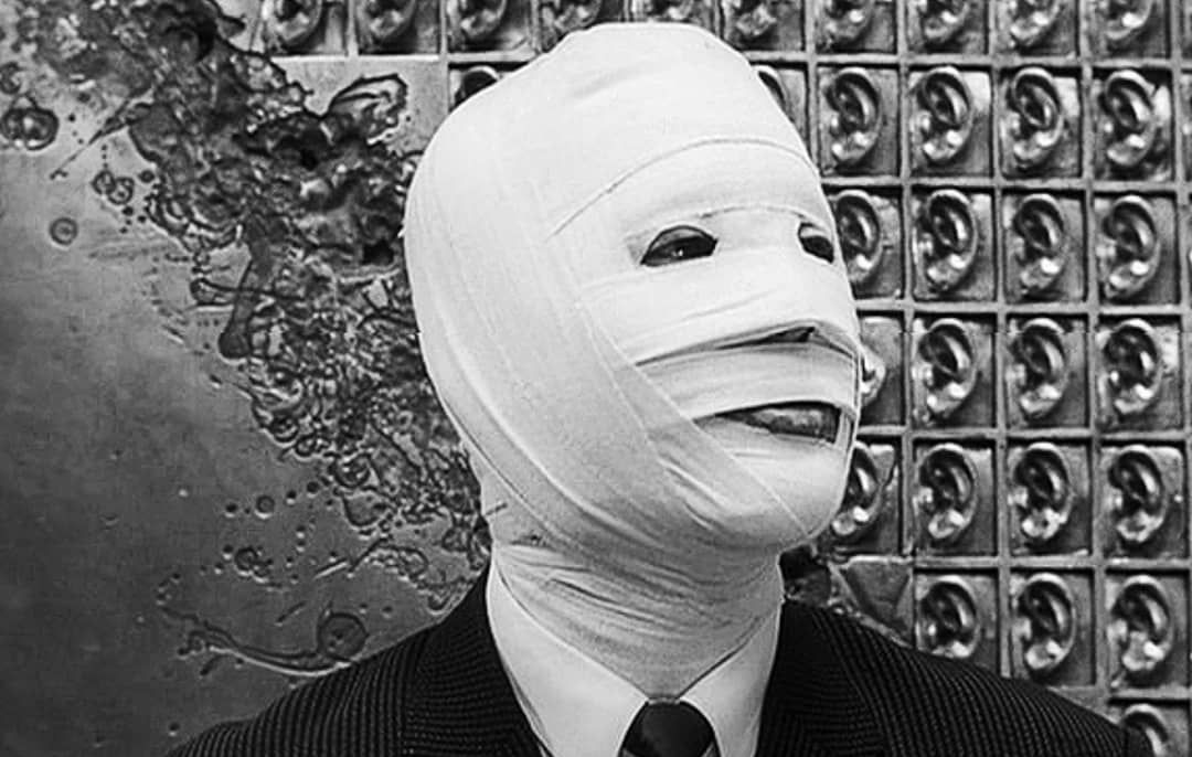 A man stands in front of a wall textured with ear carvings with his face fully wrapped in white bandages.