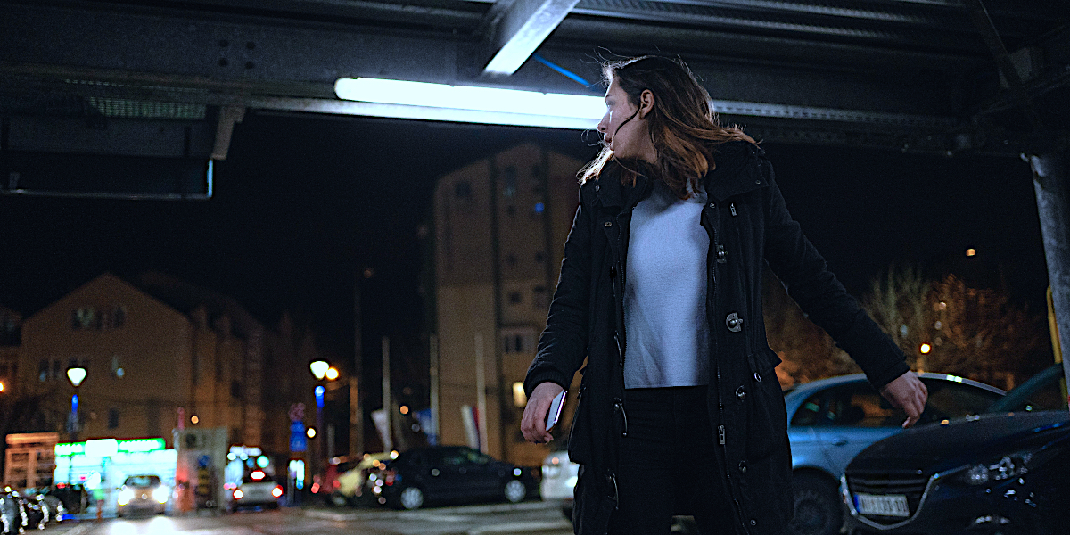 A woman looks over her shoulder in fear under a dark bridge with a glaring overhead light