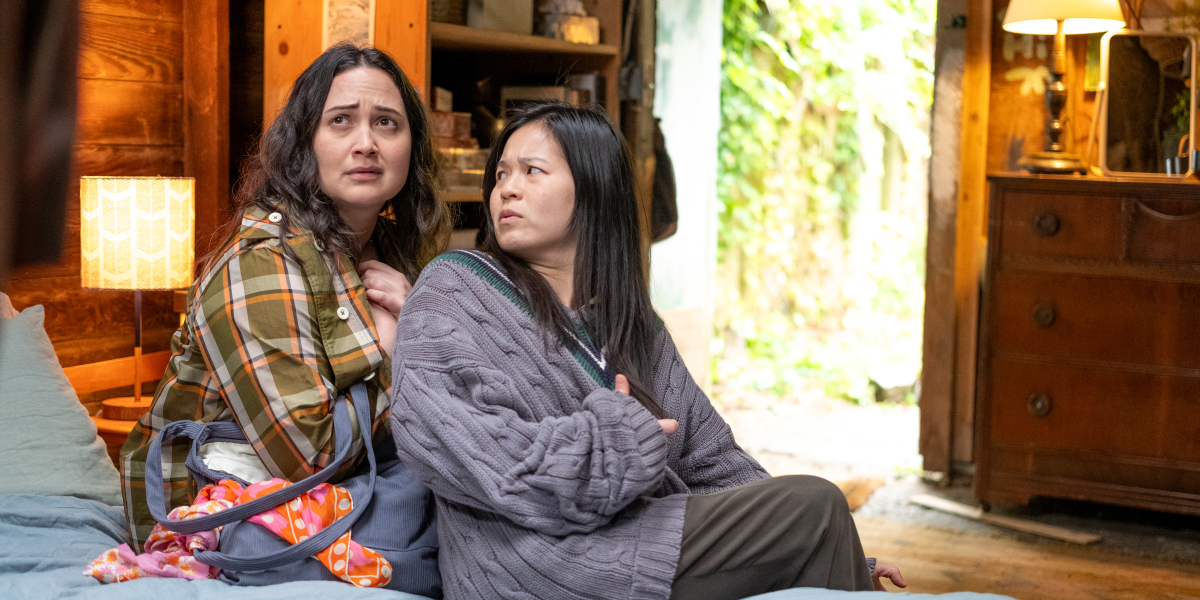 Lily Gladstone and Kelly Marie Tran look concerned sitting next to each other on a bed in flannel and sweaters.