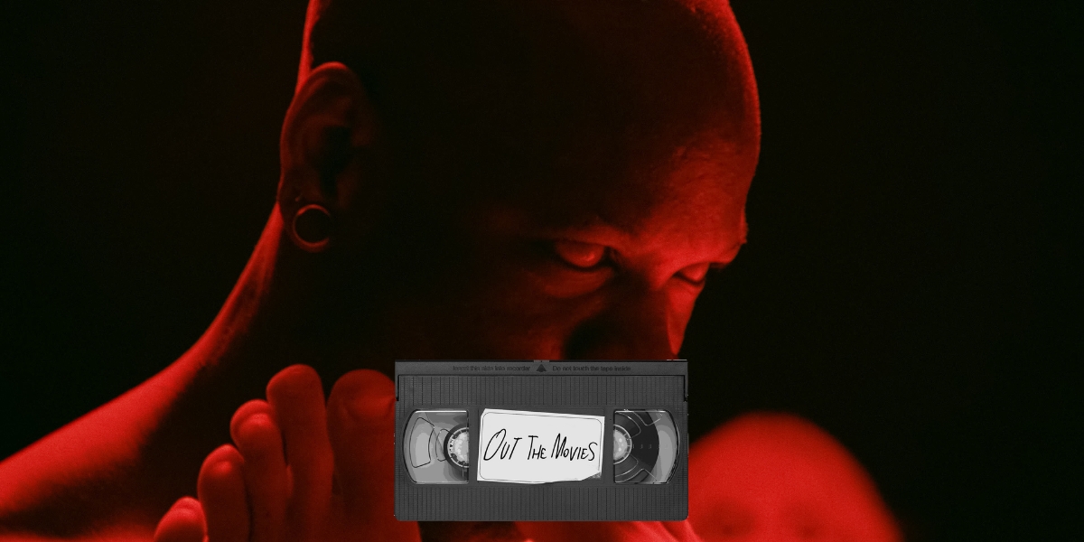Bishop Black awash in red light bites a toe in The Visitor. The Out the Movies logo at the bottom of the frame.