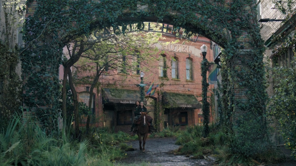 The Last of Us 204: Ellie and Dina ride on horseback through the gates of Capitol Hill, pride flags in the background