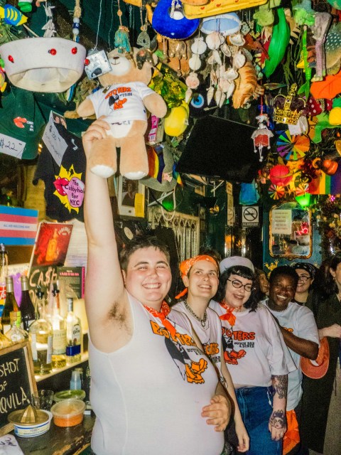 several people, including members of the Booters organizing team, all wearing Booters apparel. The person in the front is holding up a teddy bear with a Booters shirt. They're in a gay bar in NYC with objects hanging from the ceiling.