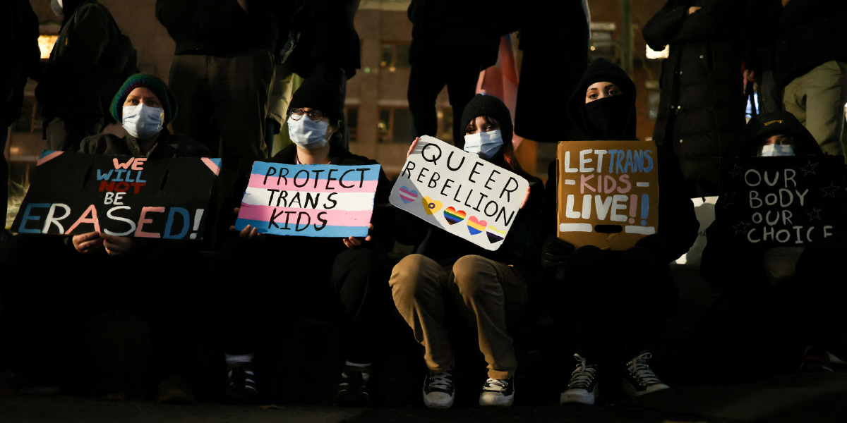 A row of protestors at night wearing masks and hoodies holding up signs that say protect trans kids and queer rebellion