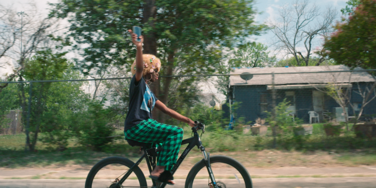 a still from Just Kids featuring a kid on a bike holding up a phone